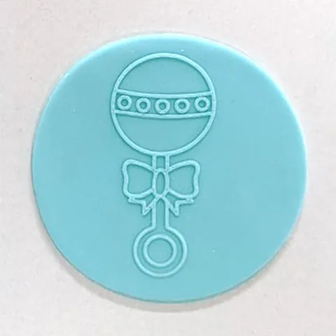 A light blue circularteal fondant disc featuring an embossed outline of a rattle with a bow, shown on a plain off-white background.