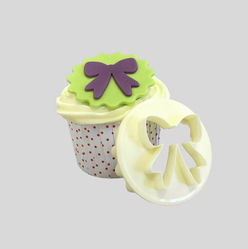 A cupcake-shaped craft cutter shown on a plain light gray background. The cutter features a white liner with red polka dots, piped cream frosting, a green scalloped top, and a purple bow decoration, with a pale yellow circular lid leaning against it.
