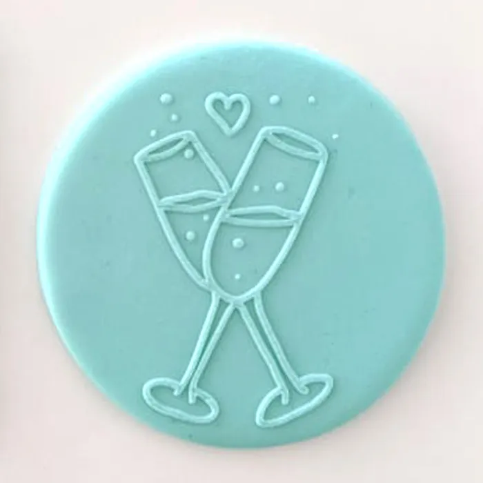 A round light blue plaque of fondant featuring a raised embossed design of two clinking champagne glasses, a small heart, and small dots on a pale pink background.