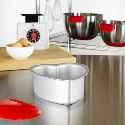 A heart-shaped aluminium cake pan sits on a brushed metal countertop, with a red silicone bowl scraper placed in front. In the background, a white bowl of eggs, a scale, and two metal mixing bowls with red silicone accents are visible on a wooden board and wire shelf.