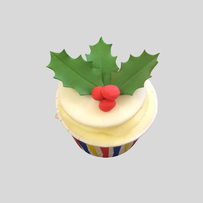 A decorated cupcake topped with white frosting, a green holly leaf decoration, and three red berries, set against a solid gray background.