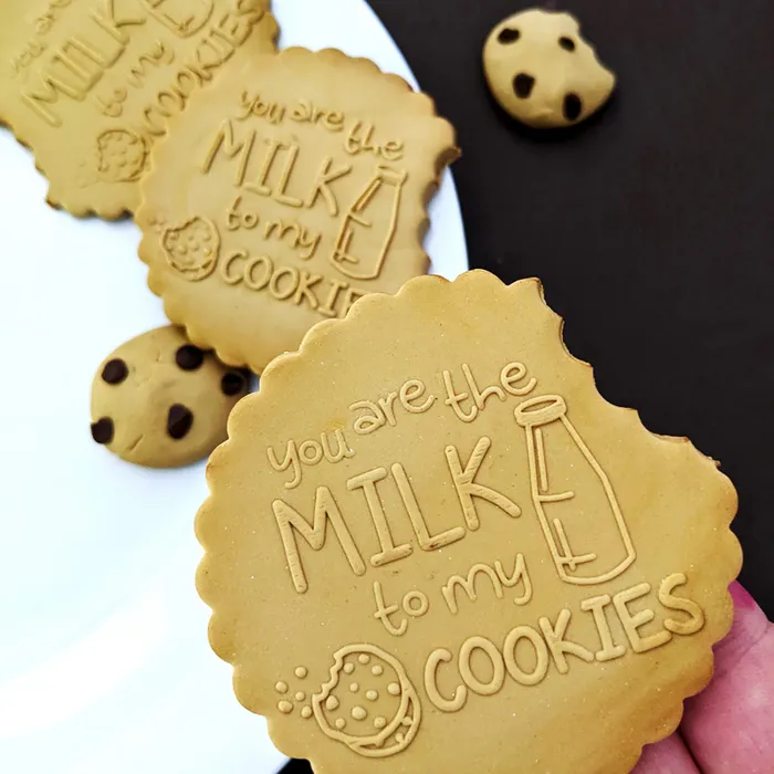 A close-up of scalloped sugar cookies baked on a white plate, with one large cookie in the foreground reading 'you are the MILK to my COOKIES' and embossed illustrations. A hand is visible holding the cookie, and a few chocolate chip-style cookies rest on a dark surface nearby.
