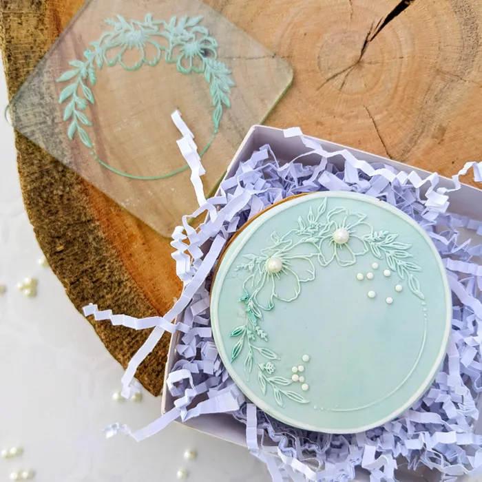 A clear debosser with a floral round frame design, displayedwith a finished cookie, used for imprinting on fondant or gumpaste icing.