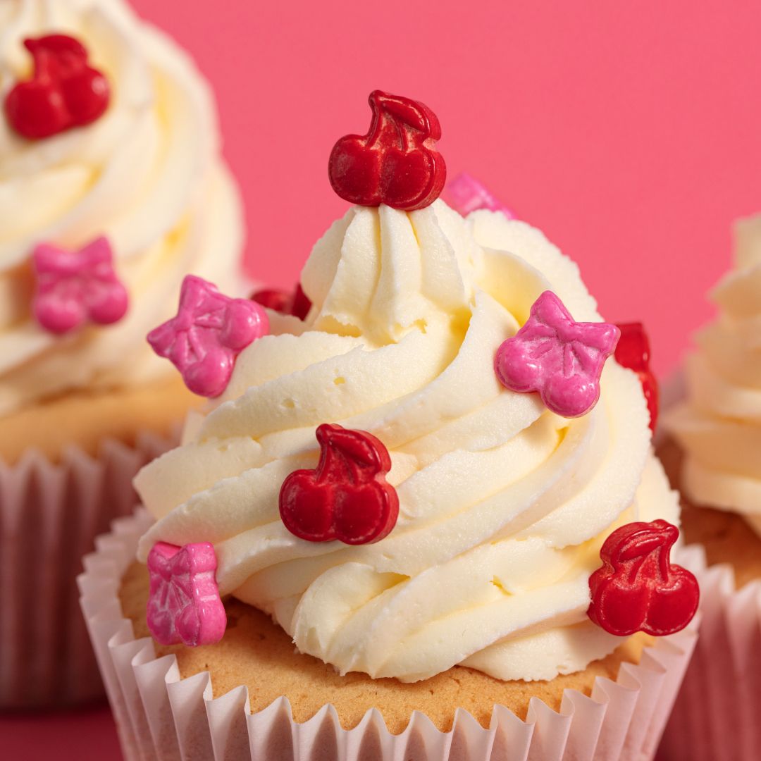 Cupcake with white frosting, red cherries, and pink bow decorations on a pink background