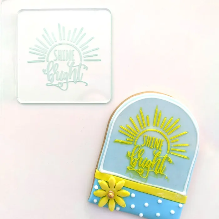 A clear embosser with the text 'Shine Bright' on a white background. With a finished cookie with a yellow and blue background.