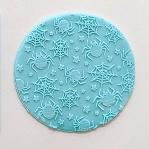 A round turquoise blue fondant icing decorative plaque featuring a repeating pattern of embossed spiders and spiderwebs on a light background.