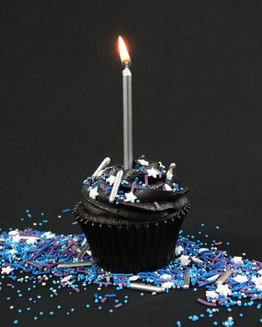 A black frosted cupcake with a single lit silver candle, topped with white, blue, and purple sprinkles, set against a dark background with sprinkles scattered around it.