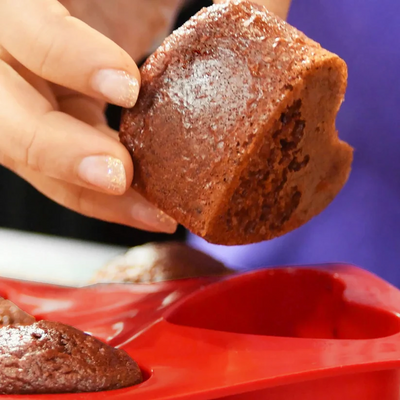 Hand lifting a freshly baked chocolate muffin from a red heart silicone mini cake mold against a purple background