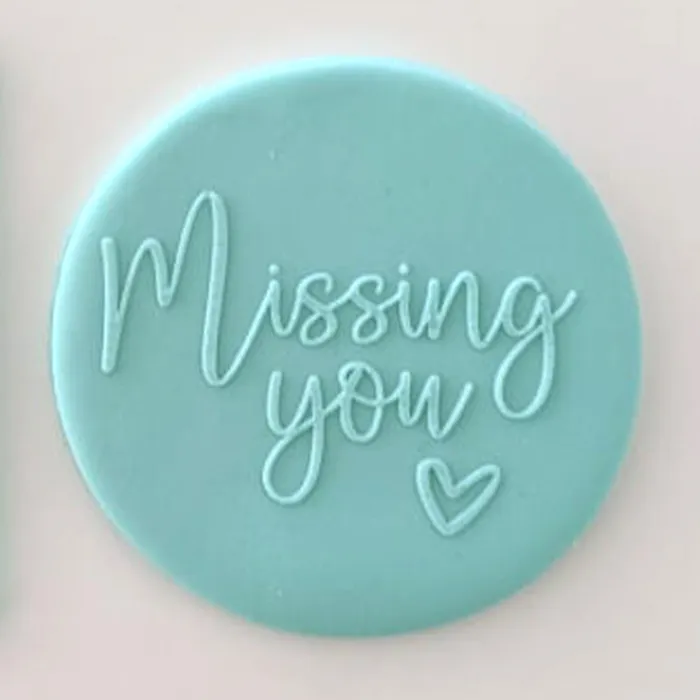 A round light blue fondant plauqe with the words 'Missing you' and a small heart in matching light blue icing, shown on a plain light background.