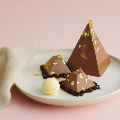 Chocolate pyramid shaped desserts with gold flakes on a white plate against a pink background