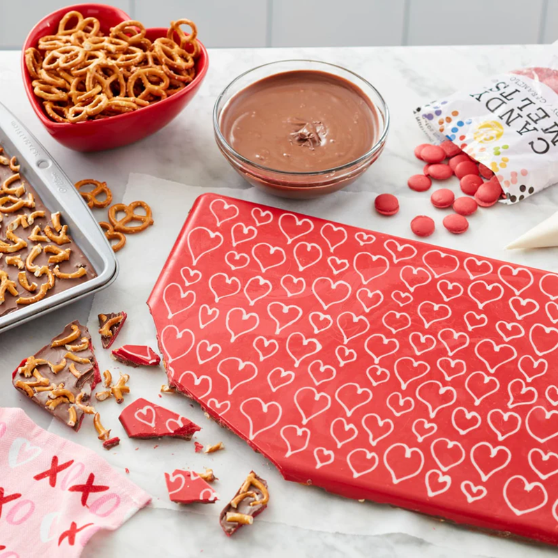 Chocolate bark with pretzels, a bowl of chocolate sauce, and a bag of candy melts on a marble surface.