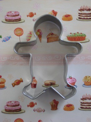 A stainless steel cookie cutter in the shape of a gingerbread man, placed on a surface with printed gingerbread cookies and other sweets.