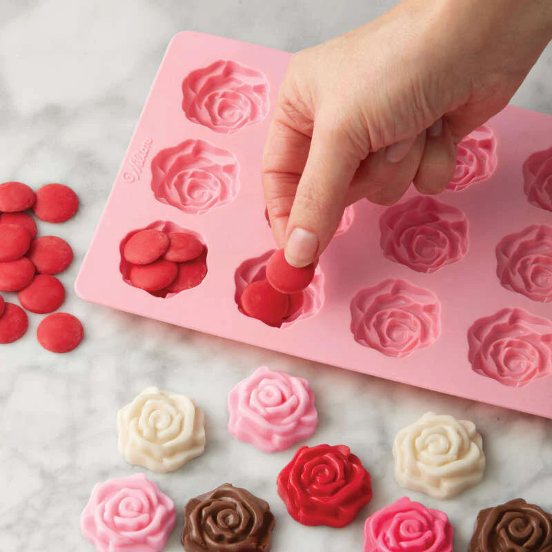 a pink silicone chocolate mould of roses. With a hand filling the cavities with coloured chocolate buttons in red pink, white and brown