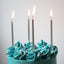 A close-up of a blue frosted cake topped with piped swirls, sprinkles, and five tall silver birthday candles with lit flames, set against a plain light gray background.