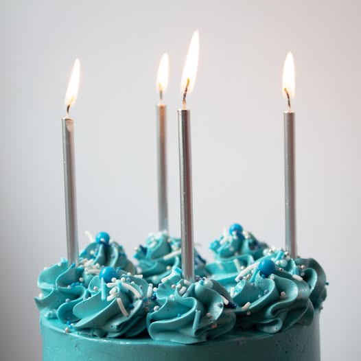 A close-up of a blue frosted cake topped with piped swirls, sprinkles, and five tall silver birthday candles with lit flames, set against a plain light gray background.