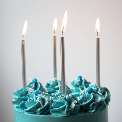 A close-up of a blue frosted cake topped with piped swirls, sprinkles, and five tall silver birthday candles with lit flames, set against a plain light gray background.