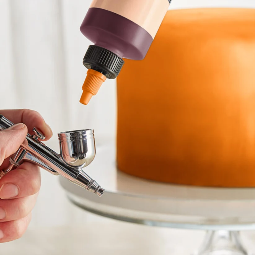 A close-up of a hand holding an airbrush tool while a squeeze bottle with a dark purple cap and orange nozzle dispenses liquid onto a round orange object on a glass stand.