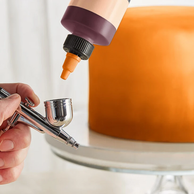A close-up of a hand holding an airbrush tool while a squeeze bottle with a dark purple cap and orange nozzle dispenses liquid onto a round orange object on a glass stand.