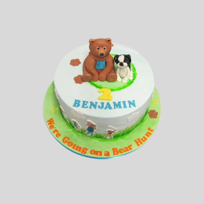 A custom round birthday cake decorated with a white top, a brown teddy bear and a black-and-white dog on top, a yellow number 2, and the name 'BENJAMIN' in blue icing. The cake sits on a green base with orange text reading 'We're Going on a Bear Hunt' and small icing characters.
