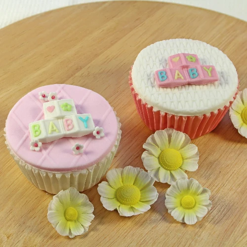 Two decorated cupcakes on a wooden surface, one with pink frosting and white fondant lettering that reads BABY, and the other with white frosting and pink fondant lettering that reads BABY. Four white and yellow flower decorations are placed in front.