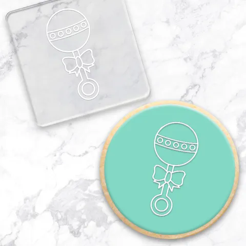 Circular cookie with a baby rattle design on a marble surface. Along with the debossing tool used to create it