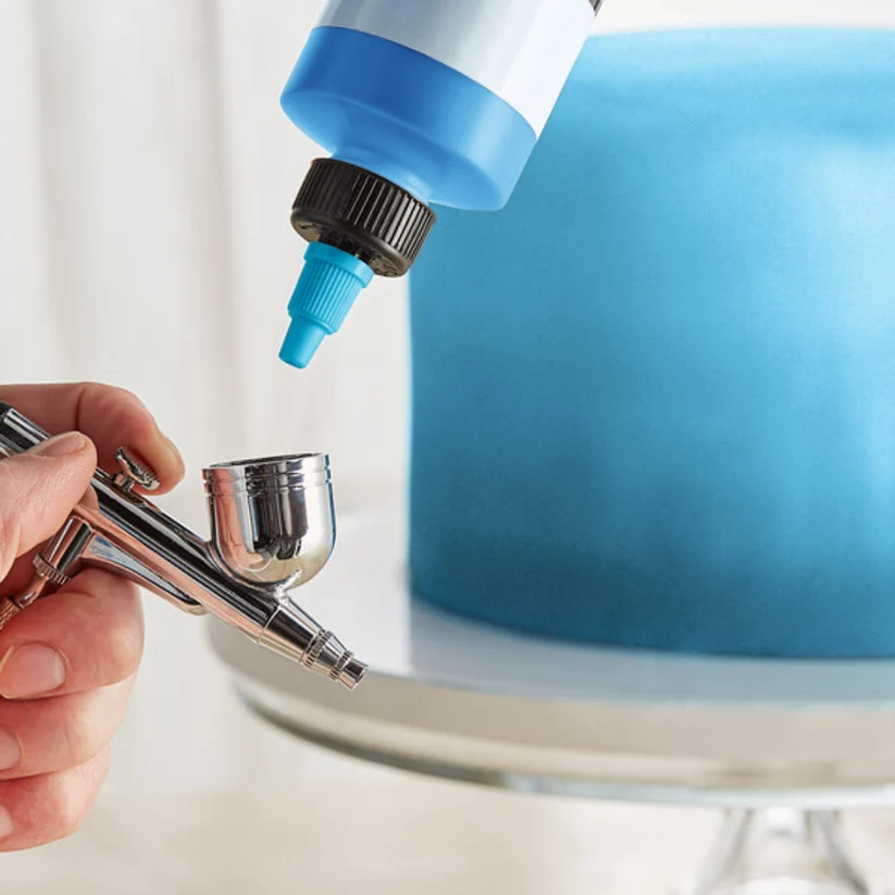 A close-up of a hand holding a silver airbrush tool while a blue squeeze bottle is positioned above it, with a blue frosted cake on a white cake stand in the background.