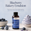 A bottle of LorAnn Better Than Extracts Blueberry Bakery Emulsion stands centered on a light surface, with blueberries in a bowl and around it and a blueberry-flavored dessert swirl to the right. Large text at the top reads 'Blueberry Bakery Emulsion' with a tagline below and the LorAnn brand visible on the bottle.