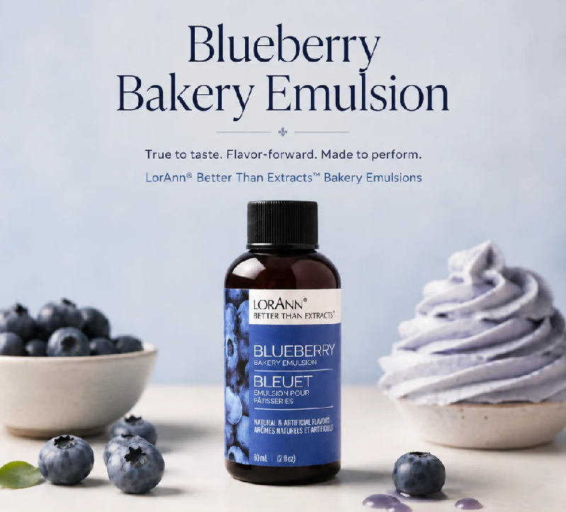 A bottle of LorAnn Better Than Extracts Blueberry Bakery Emulsion stands centered on a light surface, with blueberries in a bowl and around it and a blueberry-flavored dessert swirl to the right. Large text at the top reads 'Blueberry Bakery Emulsion' with a tagline below and the LorAnn brand visible on the bottle.