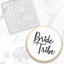 A square debosser with the phrase “Bride Tribe” in script lettering, in clear acrylic. And a cookie with white with black lettering, as an example of how to use it. both on a white marble surface.