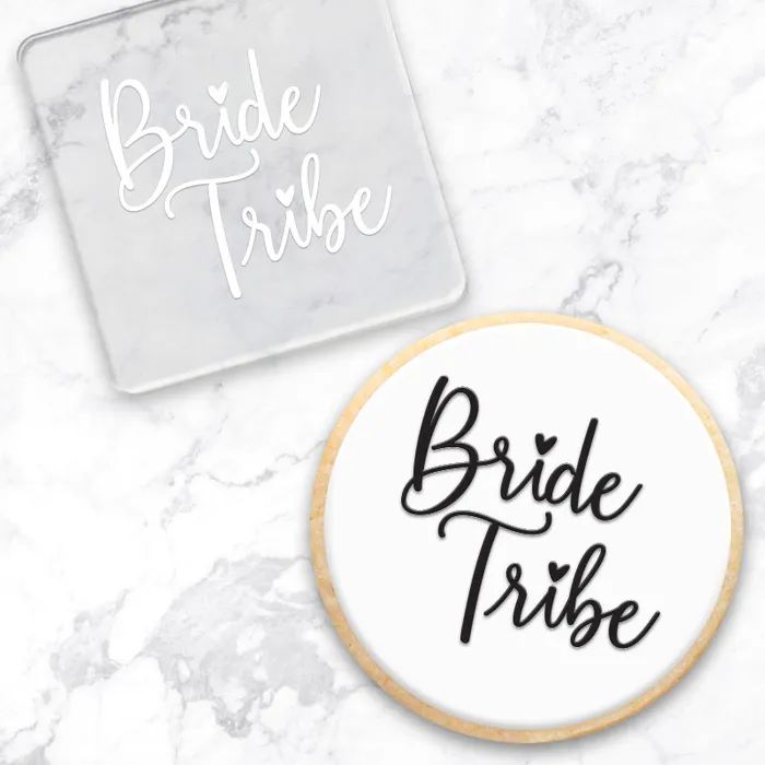 A square debosser with the phrase “Bride Tribe” in script lettering, in clear acrylic. And a cookie with white with black lettering, as an example of how to use it. both on a white marble surface.