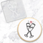 A cookie with a black and red champagne toast illustration, shown alongside a clear square debosser with a matching champagne toast design on a white marble surface.