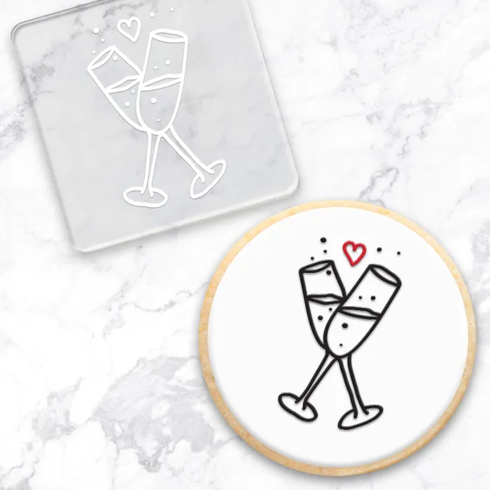 A cookie with a black and red champagne toast illustration, shown alongside a clear square debosser with a matching champagne toast design on a white marble surface.