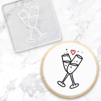 A cookie with a black and red champagne toast illustration, shown alongside a clear square debosser with a matching champagne toast design on a white marble surface.