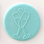 A round light blue plaque of fondant featuring a raised embossed design of two clinking champagne glasses, a small heart, and small dots on a pale pink background.