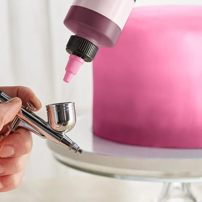 A close-up of an airbrush tool being used to apply pink icing or paint to a round cake on a stand, with a bottle of pink icing held above the tool.