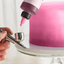 A close-up of an airbrush tool being used to apply pink icing or paint to a round cake on a stand, with a bottle of pink icing held above the tool.