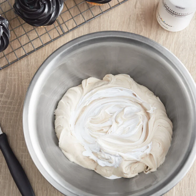 A top-down view of a stainless steel bowl containing a thick, swirled cream or frosting mixture, set on a light wood surface with baking tools nearby.