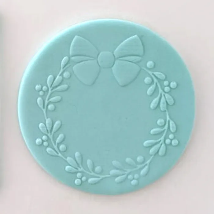 A round light blue fondant decorative plaque featuring an embossed floral wreath with a bow at the top, shown on a plain light background.