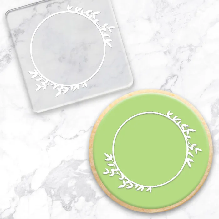 Clear acrylic debosser with circle of leaves frame. Along with a finished green cookie, showing example of use