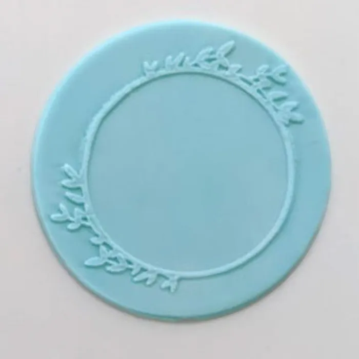 A circular light blue fondant plaque  with a decorative leafy wreath border, shown on a plain off-white background.