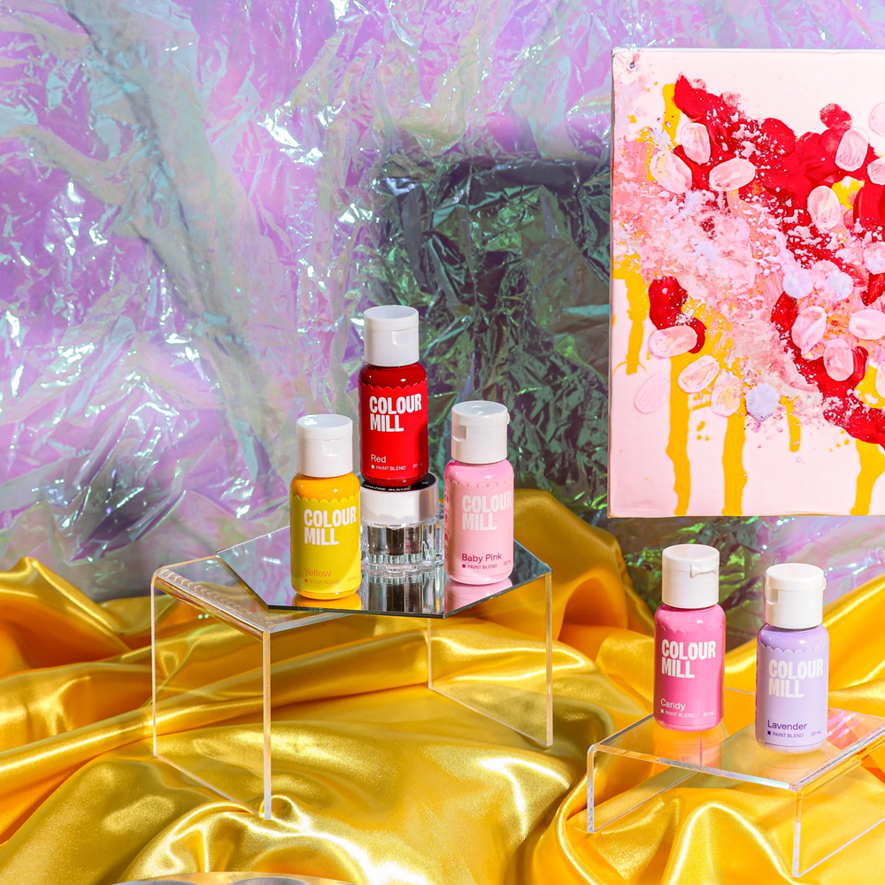 Colorful bottles of Colour Mill paint blend on a reflective surface with a colorful abstract painting in the background.