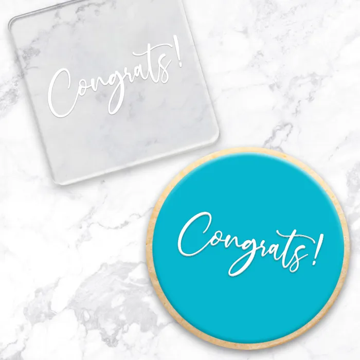 A clear square acrylic debosser with cursive 'Congrats!' text, shown alongside a round turquoise cookie with the same white script on a white marble surface.