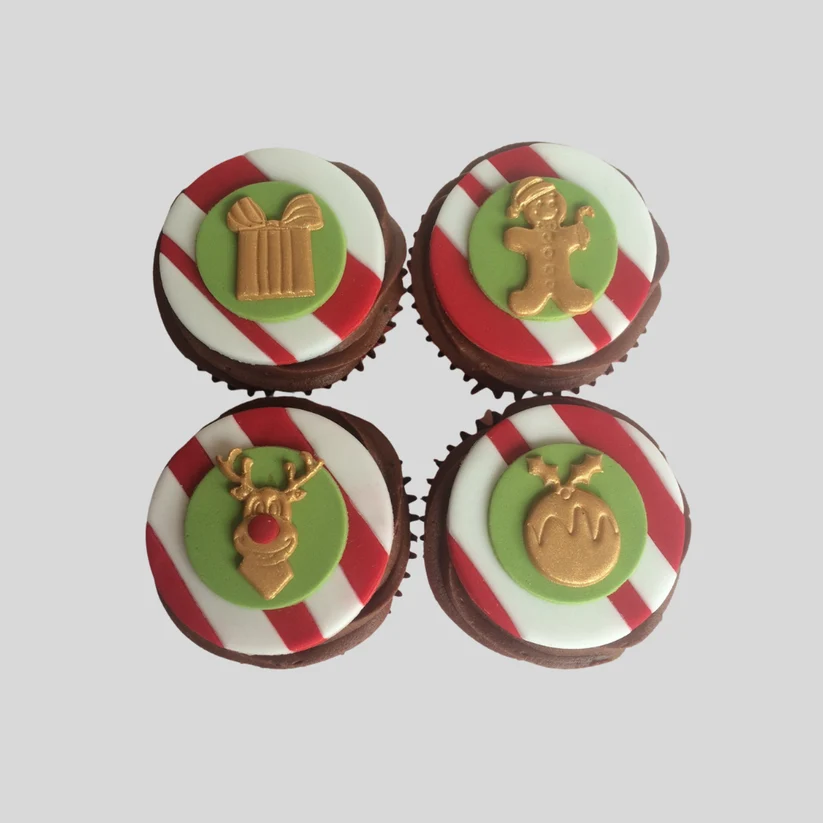 Four decorated chocolate cupcakes arranged in a 2x2 grid on a light gray background. Each cupcake has a red and white striped base, a green circular topper, and a gold holiday-themed decoration: a gift box, gingerbread man, reindeer, and mistletoe.