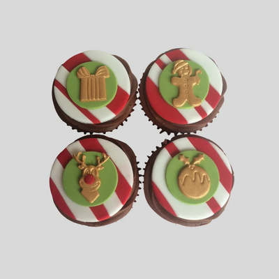 Four decorated chocolate cupcakes arranged in a 2x2 grid on a light gray background. Each cupcake has a red and white striped base, a green circular topper, and a gold holiday-themed decoration: a gift box, gingerbread man, reindeer, and mistletoe.