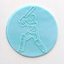A circular light blue plaque of fondant icing featuring an embossed white outline of a cricket player holding a bat, shown on a plain off-white background.