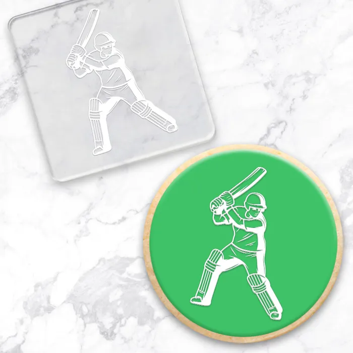 A fondant covered cookie with a bright green center and a white line-art graphic of a cricket player, shown alongside a clear acrylic debosser tool with the same design on a white marble background.