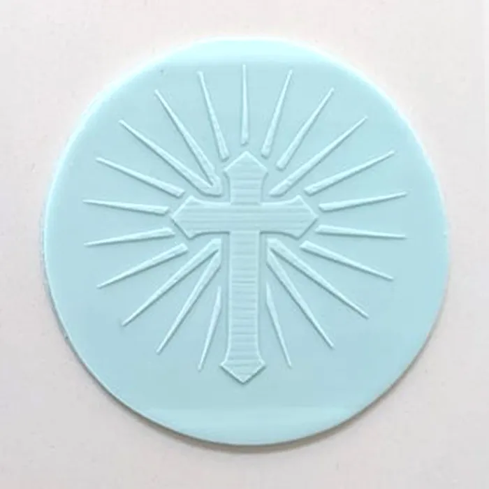A circular light blue fondant disc featuring an embossed cross with radiating lines, shown on a plain off-white background.