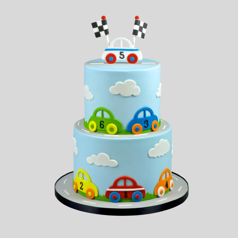 A two-tier birthday cake decorated with a light blue sky theme, featuring white clouds and colorful fondant race cars in green, blue, yellow, red, and orange. The top of the cake has a small white car with black-and-white checkered flags on top, all displayed on a black and gray circular cake board against a solid gray background.