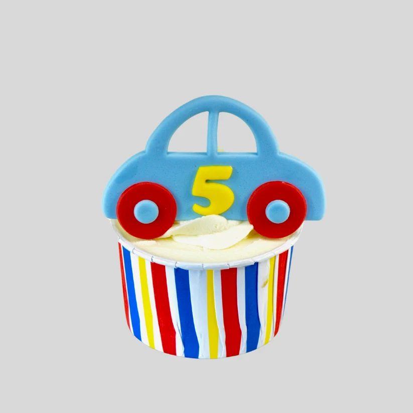 A cupcake decoration shaped like a blue car with red wheels and a yellow number 5 on top, placed on a striped cupcake liner with white frosting against a light gray background.