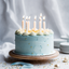 Blue cake with lit candles on a wooden stand against a light gray background